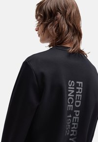 Felpa nera con collo rotondo, caratterizzata da un grande testo grigio che recita "FRED PERRY SINCE 1952" in verticale sulla schiena. Tessuto morbido e spesso.