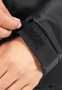 Sort vandtæt jakkeærme med velcro-lukning. Glat tekstur, enkelt design og tæt billede af en hånd, der griber fat i stoffet.