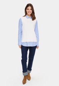Weißes ärmelloses Top über einem blauen gestreiften Hemd, kombiniert mit dunkler Jeans, die an den Bündchen umgeschlagen ist, und hellbraunen Wildleder-Ankle-Boots.