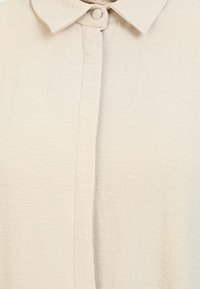 Beige blouse with a textured, lightweight fabric, featuring a classic collar and button closure down the front.