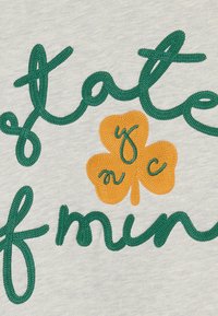 Svetlosivé látka obsahuje zelenú a oranžovú výšivku textu „State of Mind“ spolu s dizajnom štvorlístka.