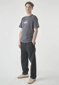 Man standing with hands in pockets wearing a dark gray graphic t-shirt, black pants, beige sneakers, and silver jewelry against a white background.