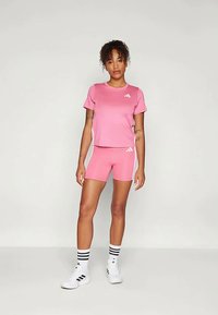 Ženska stoji v roza športnem kompletu Adidas z usklajenimi kratkimi hlačami, belimi športnimi čevlji Adidas in belimi nogavicami s črnimi črtami.