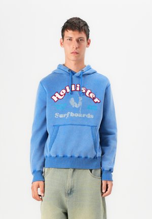 Blue hoodie featuring a front kangaroo pocket, drawstring hood, and printed "Hollister Surfboards" logo in multiple colors. Cotton blend material.