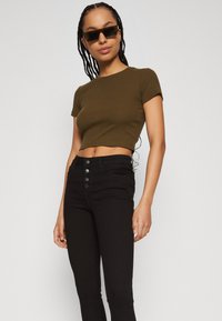 Olivenfarget ribbet crop-top kombinert med høyt liv sorte jeans med fem knapper. Modellen har på seg svarte solbriller. Glatte teksturer.