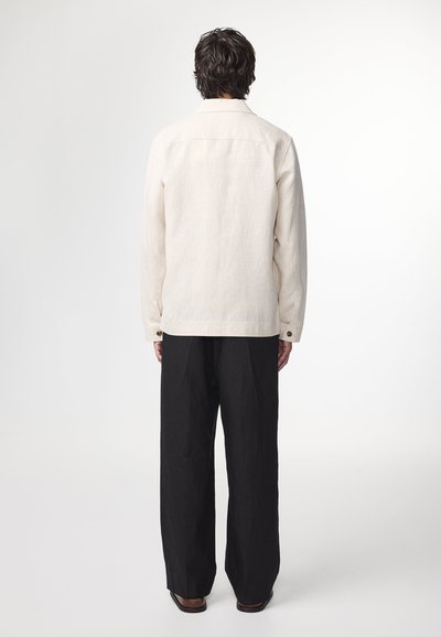 Beige linned skjorte med spidse krave, lange ærmer og knapmanchetter, parret med løstsiddende sorte bukser, set fra ryggen.