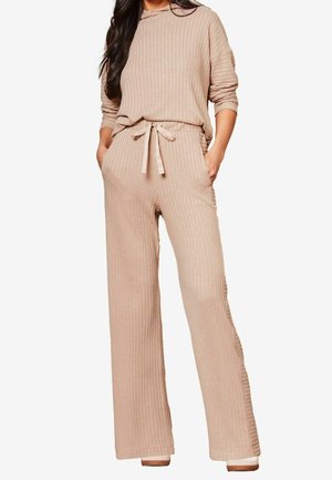 Femme portant un haut en maille côtelée beige à manches longues avec un pantalon large à taille élastiquée avec cordon, mains dans les poches, debout devant un fond blanc.