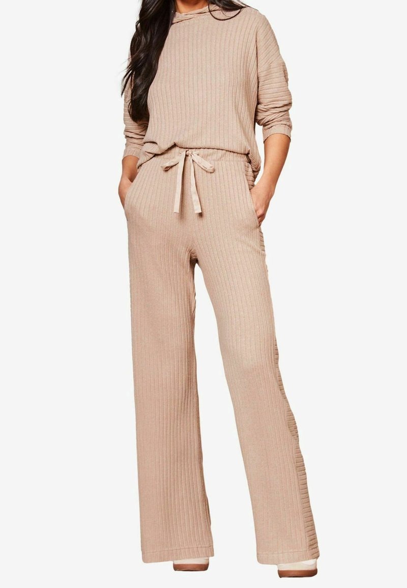 Femme portant un haut en maille côtelée beige à manches longues avec un pantalon large à taille élastiquée avec cordon, mains dans les poches, debout devant un fond blanc.