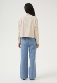 Camicia beige a righe, tagliata, con maniche lunghe e bottoni decorativi, abbinata a pantaloni in denim azzurro chiaro a gamba larga con tasche posteriori.