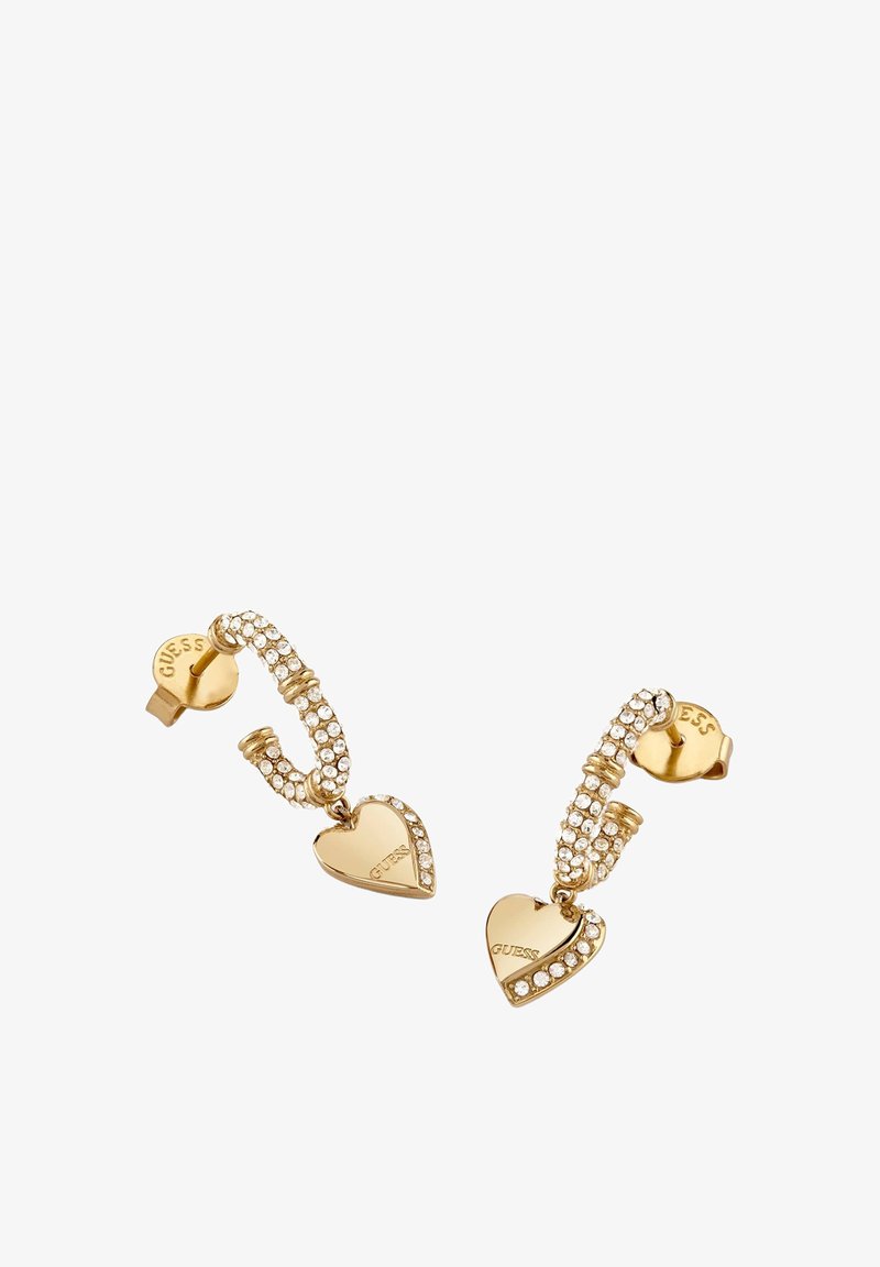 Boucles d'oreilles à clous Guess en ton or, avec un design incurvé orné de cristaux et des breloques en forme de cœur doré suspendues, gravées du mot "GUESS".