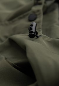 Primo piano di un tessuto verde oliva con chiusura a cordoncino nera e bottone attaccato, che mostra la trama e i dettagli delle cuciture.