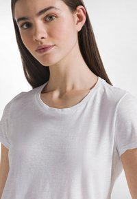 Camiseta blanca, ligera, tela con textura, cuello redondo, mangas cortas, superficie lisa, variaciones sutiles en la textura.