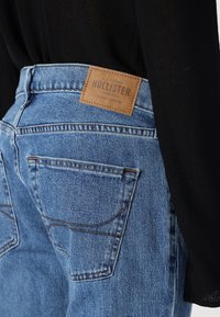 Traperice s visokim strukom u plavoj boji s kontrastnim šavovima, koje imaju smeđu kožnu oznaku s natpisom "HOLLISTER" i dvije stražnje džepove.