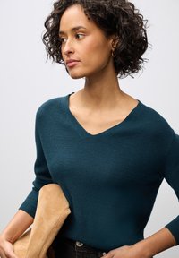 Tyrkysový, ribovaný pletený svetr s výstřihem do V a třičtvrtinovými rukávy, doplněný béžovým doplňkem, s měkkou texturou na neutrálním pozadí.