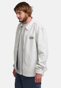 Light gray denim jacket with a structured collar, zip closure, and embroidered logo patch. Features relaxed fit and buttoned cuffs.