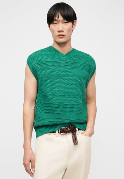 Νεαρός άνδρας που φοράει αμάνικο πράσινο πλεκτό πουλόβερ και μπεζ παντελόνι με πλεκτή καφέ ζώνη, στέκεται μπροστά από μια απλή φόντο.