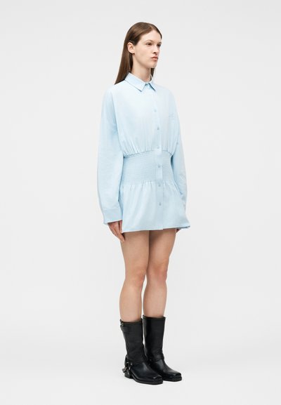 Γυναίκα που φοράει ανοιχτό μπλε φόρεμα με μακριά μανίκια και ελαστική μέση, καθώς και μαύρες δερμάτινες μπότες μέχρι τη μέση της γάμπας, στέκεται μπροστά σε απλό λευκό φόντο.