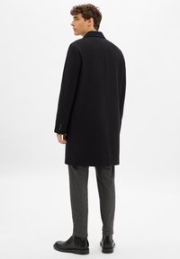 Cappotto di lana nera con un design strutturato, colletto a cappotto e maniche lunghe. Abbinato a pantaloni grigi e stivaletti neri. Linee semplici e pulite.