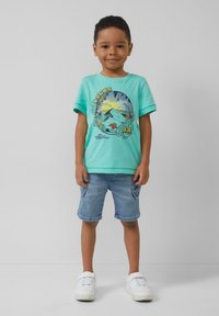 Turquoise t-shirt met korte mouwen, voorzien van een grafisch ontwerp van bergen en loof, gecombineerd met lichtdenim shorts en witte sneakers.