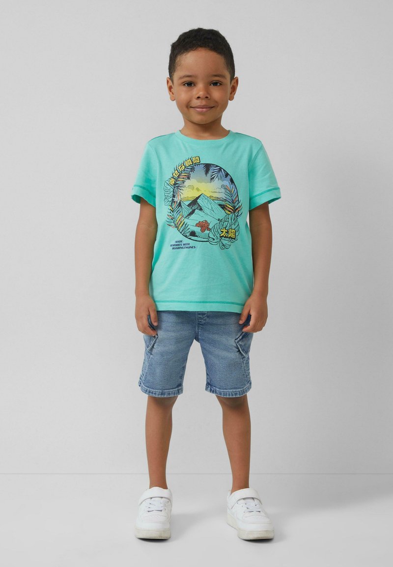 Turquoise t-shirt met korte mouwen, voorzien van een grafisch ontwerp van bergen en loof, gecombineerd met lichtdenim shorts en witte sneakers.