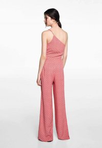 Yhden olkapään haalari vaaleanpunaisesta kankaasta, jossa on geometrinen kuvio, leveillä lahkeilla ja mukautuvalla vyötäröllä. Pehmeä rakenne ja säädettävät hihnat.