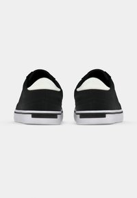 Paire de baskets basses noires avec semelles blanches vues de dos sur un fond gris clair uni.