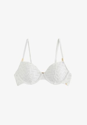 Młoda kobieta ubrana w biały teksturowany top od bikini, stojąca na zewnątrz z rozmytym naturalnym tłem.