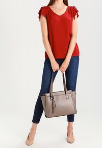 Grå læder tote taske med flettede detaljer, to håndtag og frynse. Båret med en rød top og mørke jeans, der fremhæver et afslappet look.