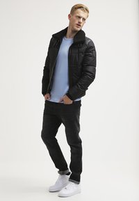 Jachetă puffer neagră cu guler texturat, pulover bleu deschis dedesubt, blugi negri și adidași albi. Poziție în picioare pe un fundal simplu.