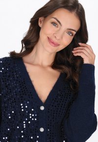 Cardigan blu navy con scollo a V, decorato con paillettes, dalla texture morbida e con bottoni argentati sul davanti. Maniche lunghe con polsini a coste.
