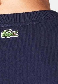 Laivastonsininen neulepusero, jossa on resorikuvio ja pyöreä kaula-aukko; vasemmalla puolella pieni vihreä krokotiililogo kirjailtuna.