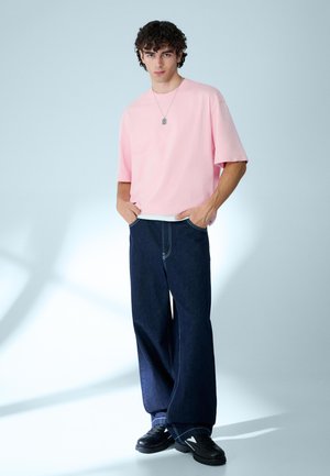 Ung mand med krøllet hår iført en løs pink T-shirt, vide mørke denimbukser, sorte sneakers og en vedhængs-halskæde, stående med hænderne i lommerne.