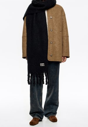 Personne portant un manteau beige boutonné, une grande écharpe noire avec l'étiquette "BIMBA Y LOLA", un jean large foncé et des chaussures en daim marron.