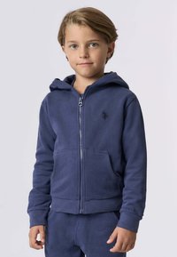 Giovane bambino con capelli castano chiaro che indossa una felpa blu navy con zip e pantaloni coordinati, in piedi davanti a uno sfondo chiaro e semplice.