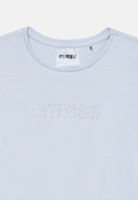 T-shirt en coton bleu clair avec col rond et manches courtes. Présente le logo "GUESS" en strass sur le devant. Étiquette de taille visible.