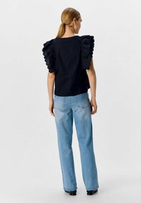 Svart topp med volangtyg på axlarna, ihopparad med ljusblå raka jeans och svarta stövlar, sedd bakifrån.