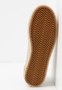 Semelle d'une chaussure avec une bande de roulement en caoutchouc marron en zigzag et du jute tissé autour du bord, positionnée à la verticale contre un coin blanc.