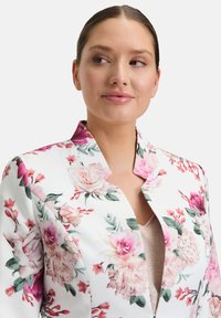 Kwiatowa marynarka na białym tle, zdobiona różowymi i czerwonymi różami, zielonymi liśćmi, o ostrym fasonie kołnierza i zapinana na zamek.