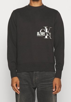 Personne portant un sweat-shirt noir Calvin Klein Jeans avec le logo CK argenté sur la poitrine et un jean gris foncé délavé.