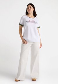 Weißes T-Shirt mit grünen Akzenten und bedrucktem Text, kombiniert mit weiten beigen Hosen und beigen Schuhen. Minimale Textur, lässige Passform.