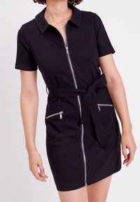 Femme portant une robe noire à manches courtes avec une fermeture éclair à l'avant, un col, une ceinture nouée à la taille et deux poches zippées près des hanches.