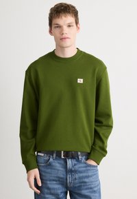 Sweatshirt verde com decote redondo, punhos canelados e um pequeno retângulo de logotipo. Usado com jeans azuis e um cinto preto, apresentando um tecido texturizado.