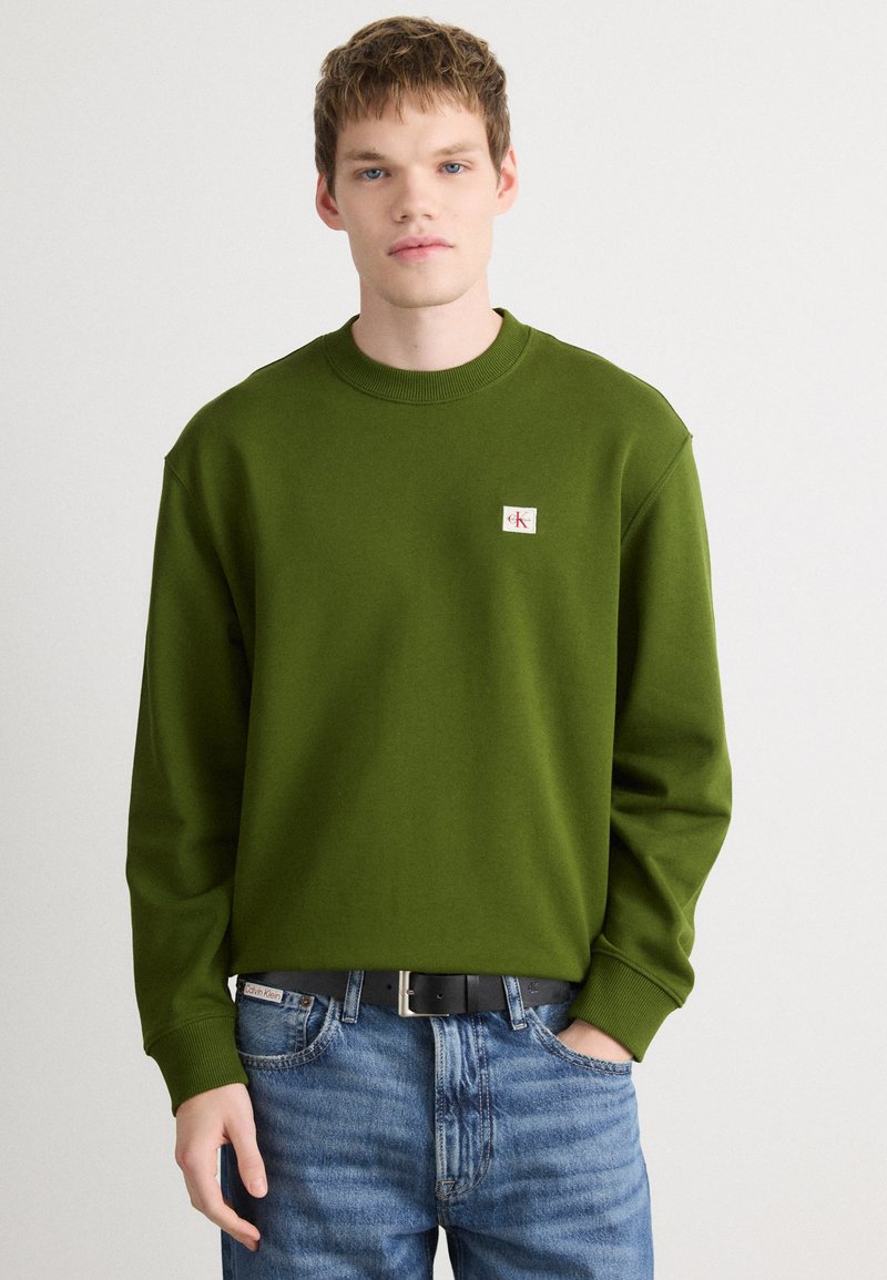 Sweatshirt verde com decote redondo, punhos canelados e um pequeno retângulo de logotipo. Usado com jeans azuis e um cinto preto, apresentando um tecido texturizado.