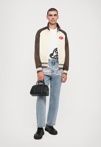 Crème en bruine jas, witte t-shirt met grafisch ontwerp, lichtblauwe jeans met roze borduursel, zwarte handtas en zwarte sneakers.