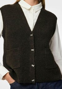 Tmavě hnědý pletený svetr s výstřihem do V, vertikálním žebrováním, dvěma předními kapsami a malými stříbrnými mašličkovými knoflíky. Nosený přes bílou košili s límečkem.
