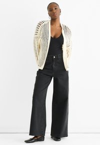 Cream-colored knitted cardigan with an open weave, worn over a black top and paired with high-waisted black wide-leg jeans. Black flats.