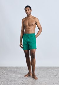 Πράσινα μαγιό με ελαστική μέση και κορδόνι, με διακοσμητικό λογότυπο. Το μοντέλο στέκεται ξυπόλητο μπροστά σε ουδέτερο φόντο.