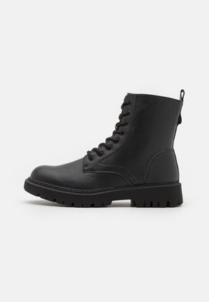 Bottines noires en cuir synthétique avec une semelle plate texturée, un bout rond et un laçage à l'avant. Petite boucle à l'arrière pour faciliter l'enfilage.