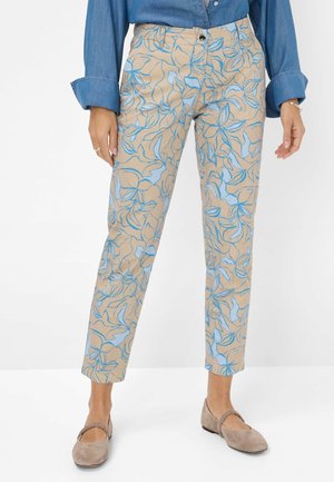 Femme portant un pantalon beige court avec un imprimé floral bleu, une chemise bleue à manches longues, et des chaussures plates beiges avec des brides élastiques.