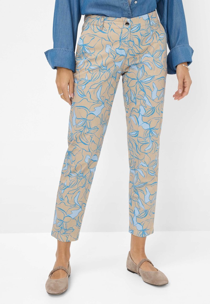 Femme portant un pantalon beige court avec un imprimé floral bleu, une chemise bleue à manches longues, et des chaussures plates beiges avec des brides élastiques.